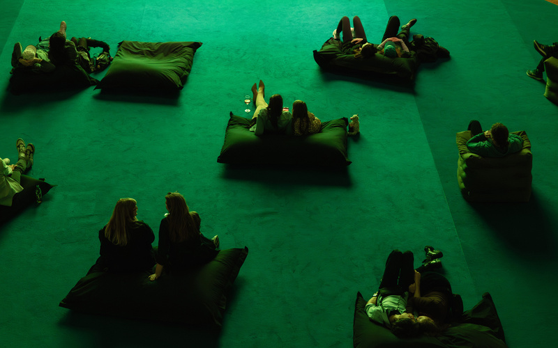 Auf dem großen Teppich sind Sitzkissen verteilt, auf denen Personen sitzen.