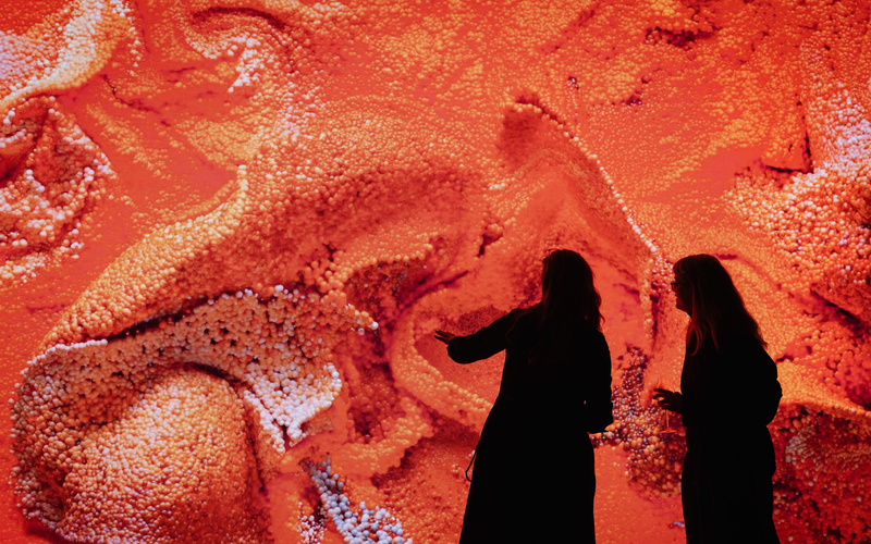 Vor einer großen Leinwand, auf der ein abstraktes Kunstwerk zu sehen ist, stehen zwei Silhouetten.