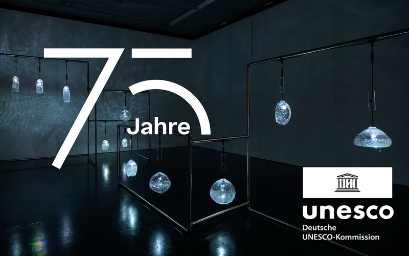 Schriftzug 75 Unesco vor einem Raum mit dem Kunstwerk von Justine Emard: Gestell mit blau leuchtenden Glasobjekten