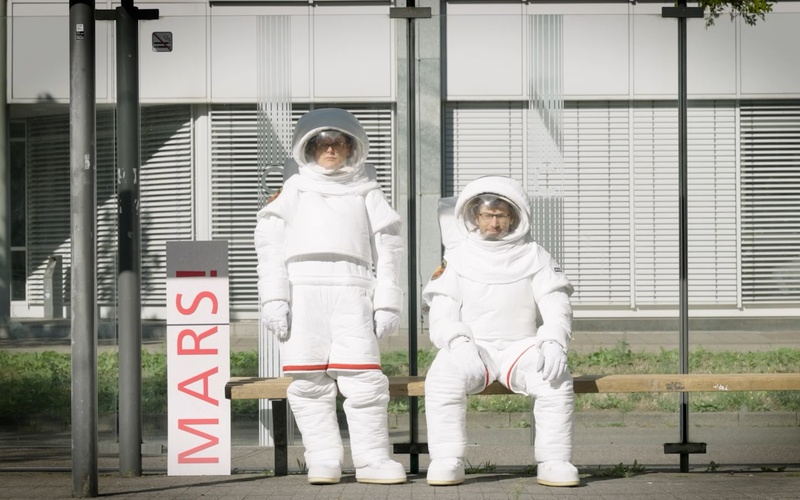 Zwei Astronaut:innen warten auf die S-Bahn.