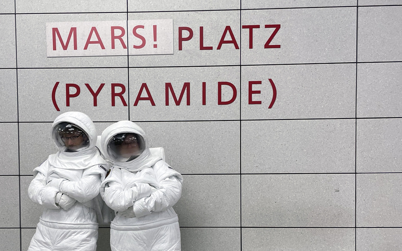Zwei Astronaut:innen in der U-Bahn vor dem Haltestellenschild MARS!platz.