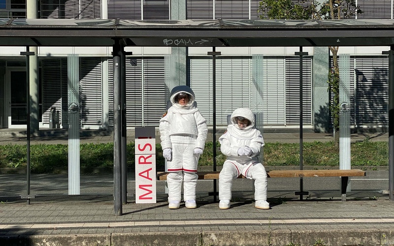 Zwei Astronaut:innen warten auf die S-Bahn.