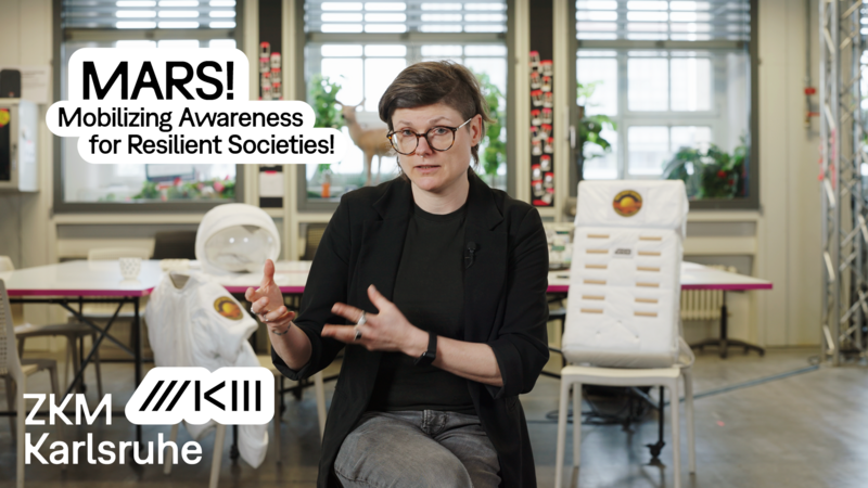 Tina Lorenz is sitting in the Hertzlab, behind her is a large table, and on the chairs are parts of an astronaut costume.