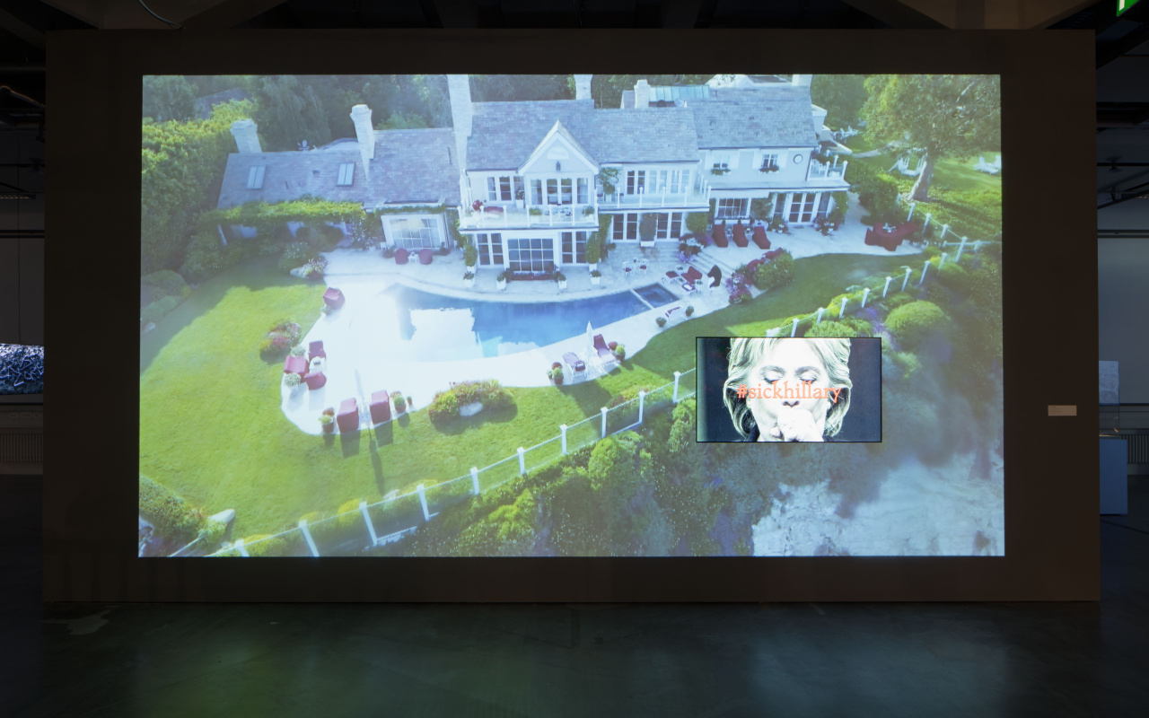 Auf einer Videoleinwand ist eine Villa mit Pool und ein Bild von Hillary Clinton unter dem »sickhillary« steht, zu sehen.