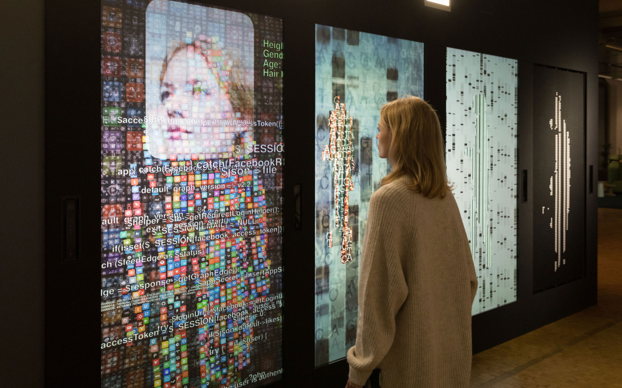 A woman stands in front of a wall with several screens and digital representations