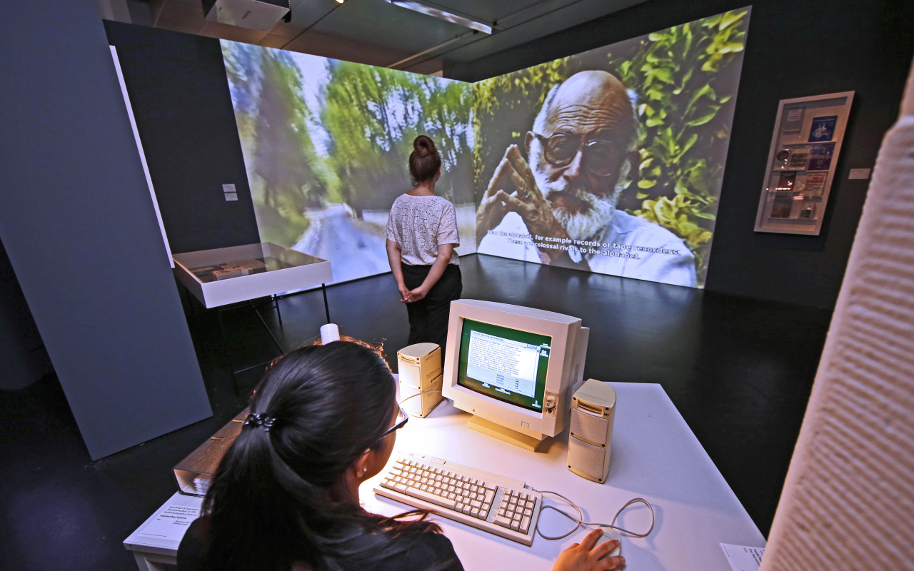 Ausstellungsansicht: ein Mädchen am Computer, im Hintergrund eine wandfüllende Videoprojektion mit Vilém Flusser