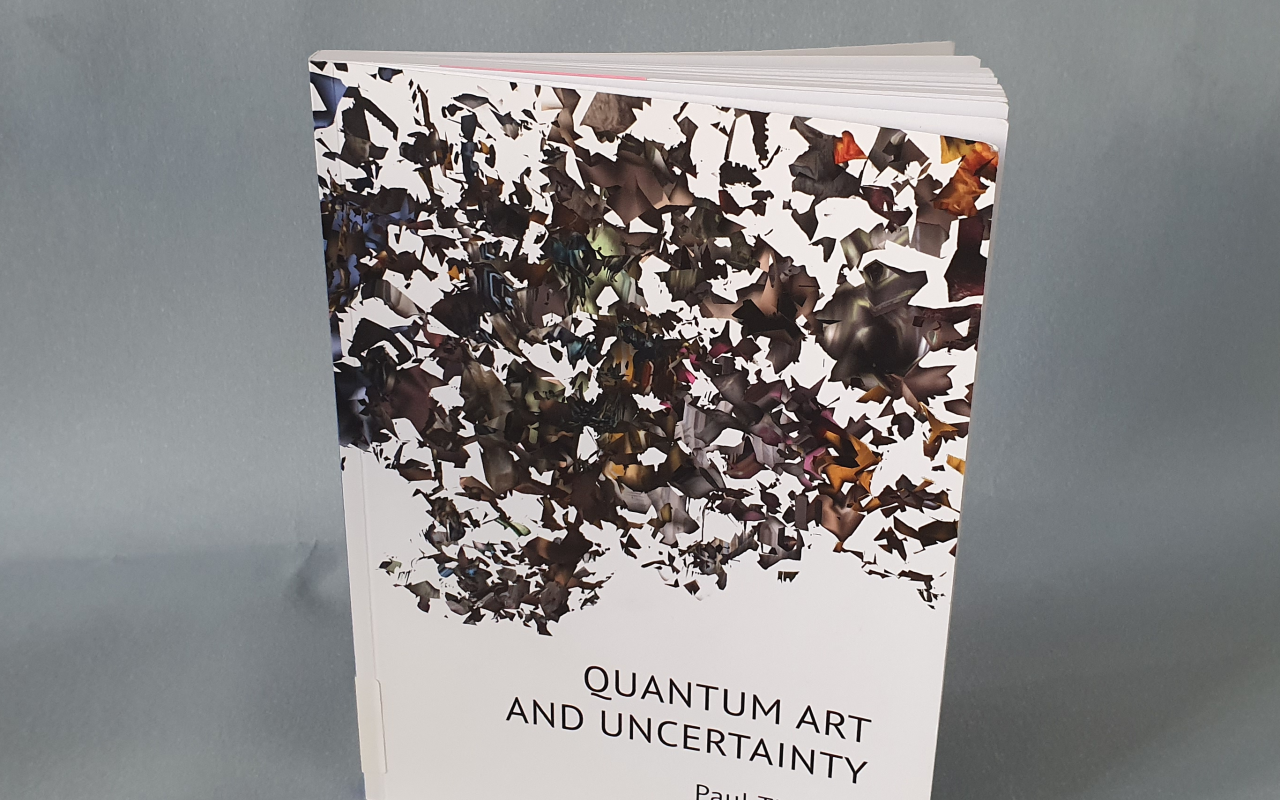 you can see the book »quantum art and uncertatinty« by paul thomas, standing, slightly open, against a blue-grey background.