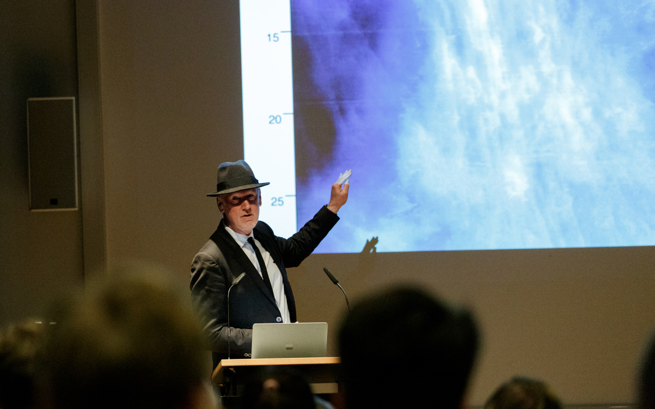 Zu sehen ist Thomas Paul, Medienkünstler und Professor in Anzug mit Hut, wie er hinter einem Rednerpult mit Laptop steht und auf die Beamer-Projektion zeigt.