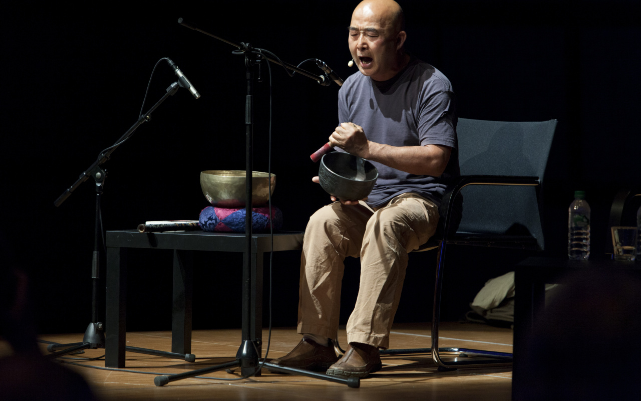 A man is sitting on a stage, making music with a  sound bowl while he is singing