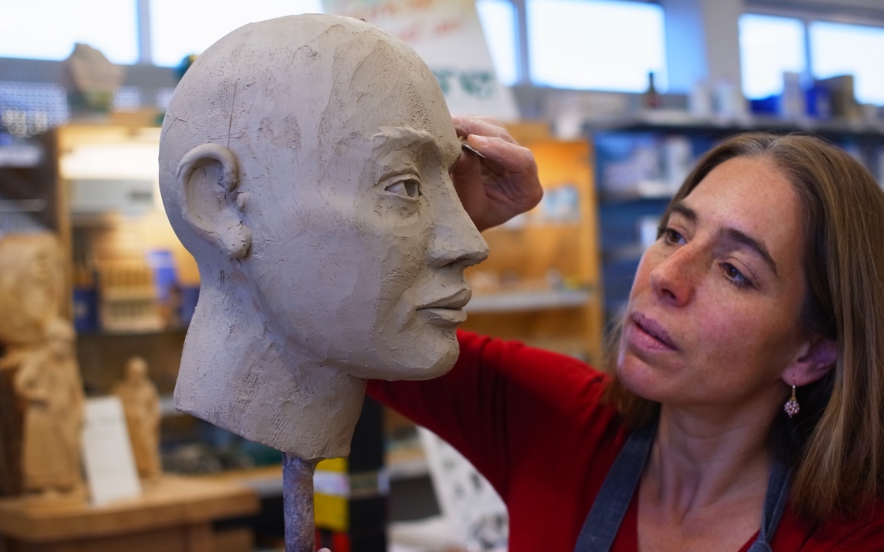 A model of a head sticking on a pole. A woman is working on it with a sculpting tool.