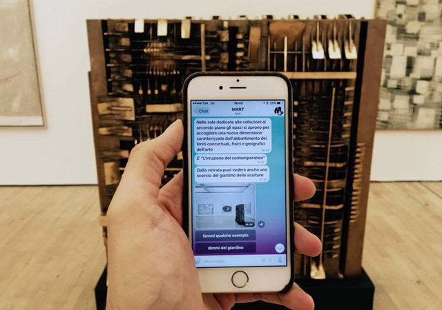 Smartphone in der Hand eines Museumsbesuchers, mit Chatnachrichten des Bots Mart, vor einem Kunstwerkim Museum 