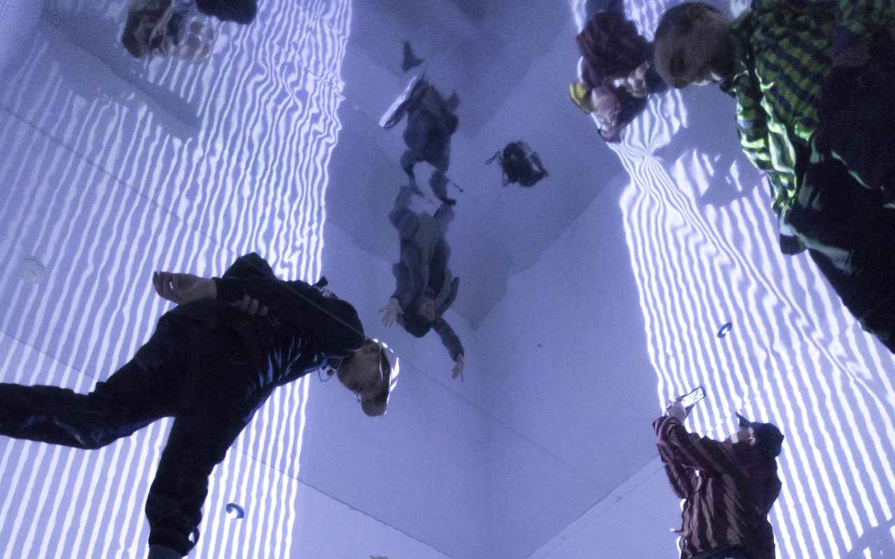 Visitors in a room with video projections on walls and ceiling and a mirrored floor.