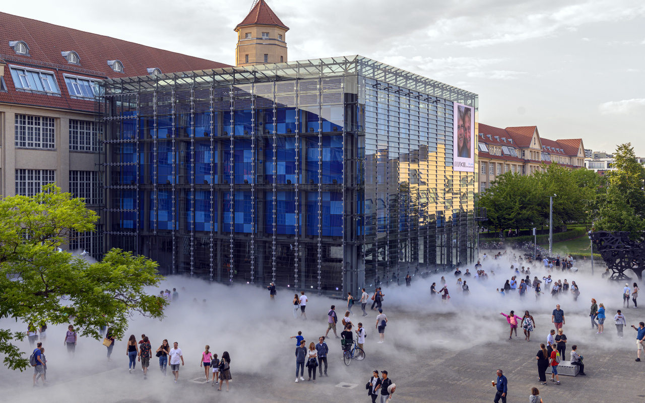 Das Foto wurde mit einer Drohne aufgenommen und zeigt den Kubus des ZKM bei Tag. Drum herum sind eine Menge Menschen versammelt die hinter der Nebelskulptur zum Teil verdeckt werden.