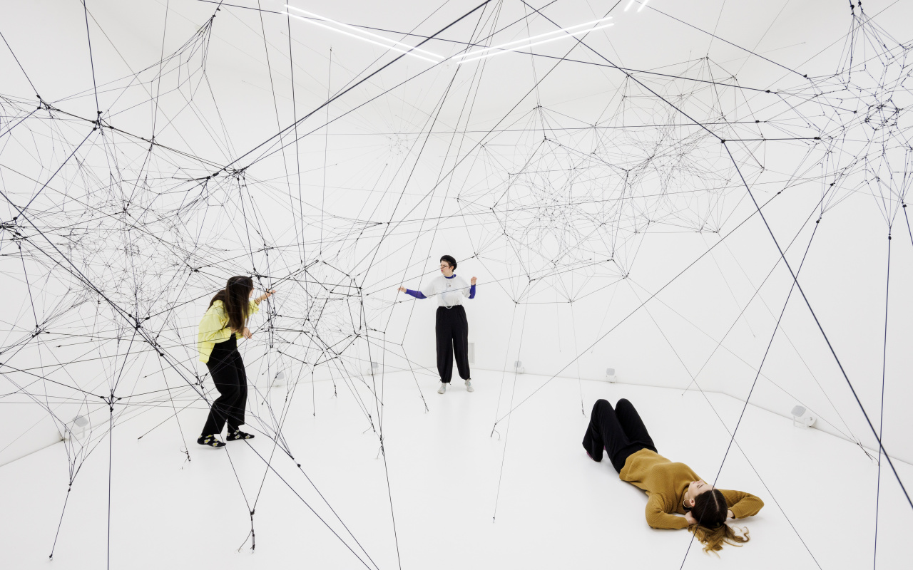 You can see a large net of threads in a white room. Several people are standing in this space and touching these threads.