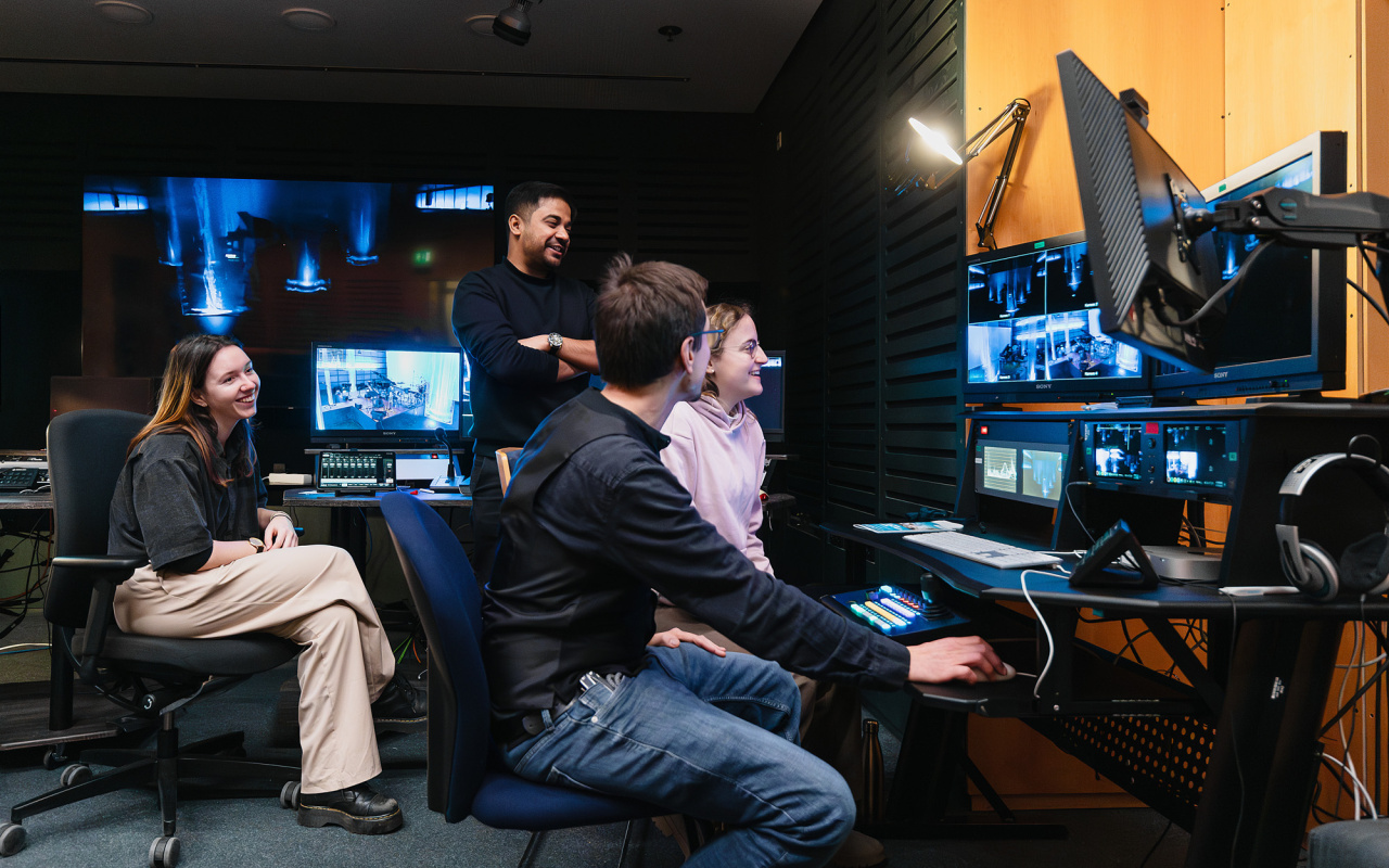 Das Bild zeigt die Mitarbeiter:innen des Videostudios im Regieraum auf Monitore blickend.