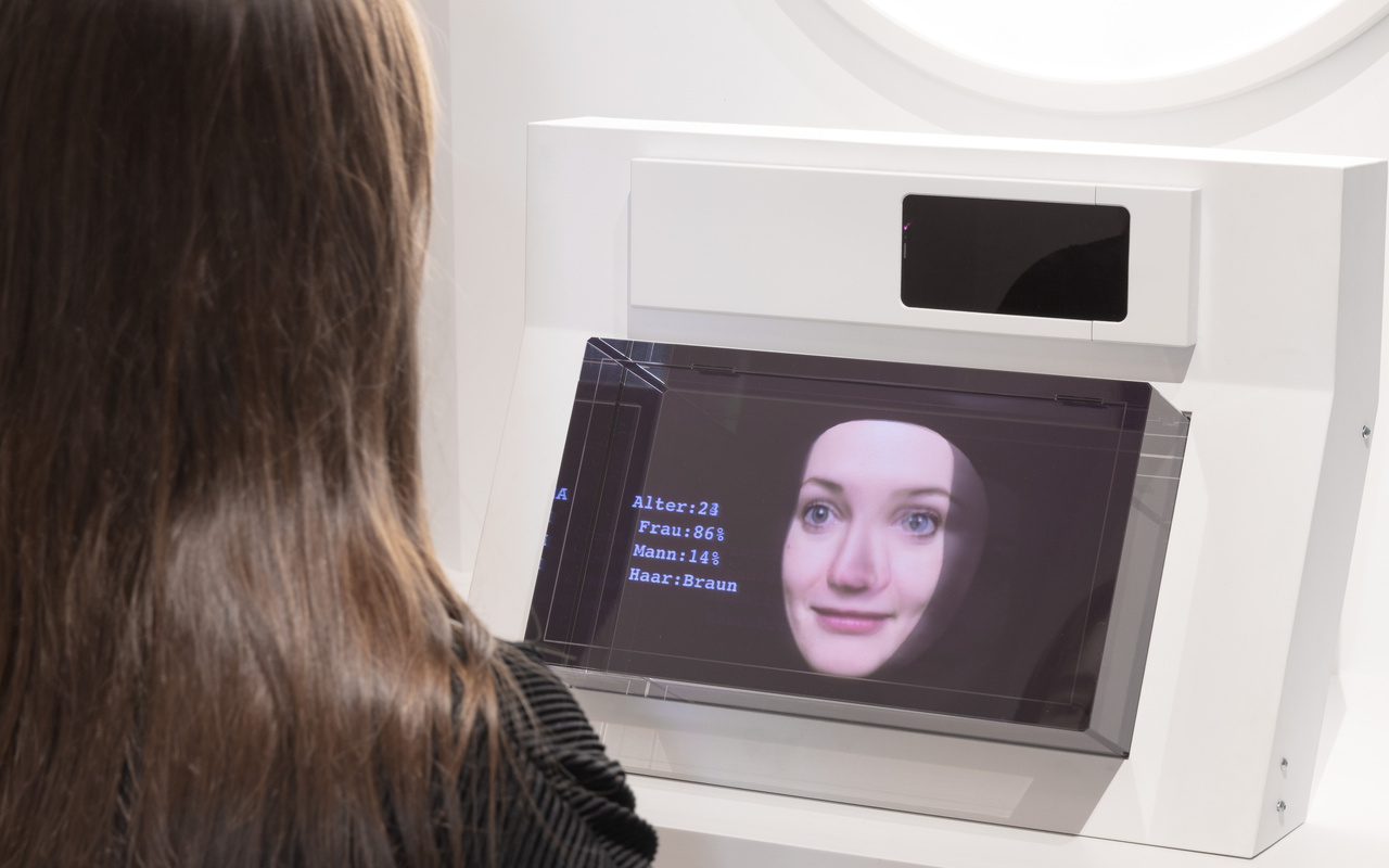 Zu sehen ist eine Besucherin. Sie sitzt mit dem Rücken gekehrt und interagiert mit der Medieninstallation. Sie spricht mir einer ihr zugewandten weiblichen künstlichen Intelligenz.