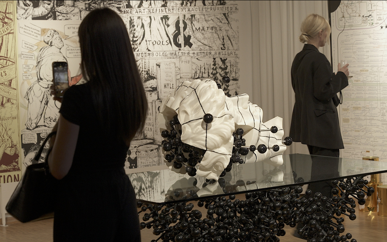 Two women walk through the exhibition. In the centre is a white sculpture of a head studded with black spheres