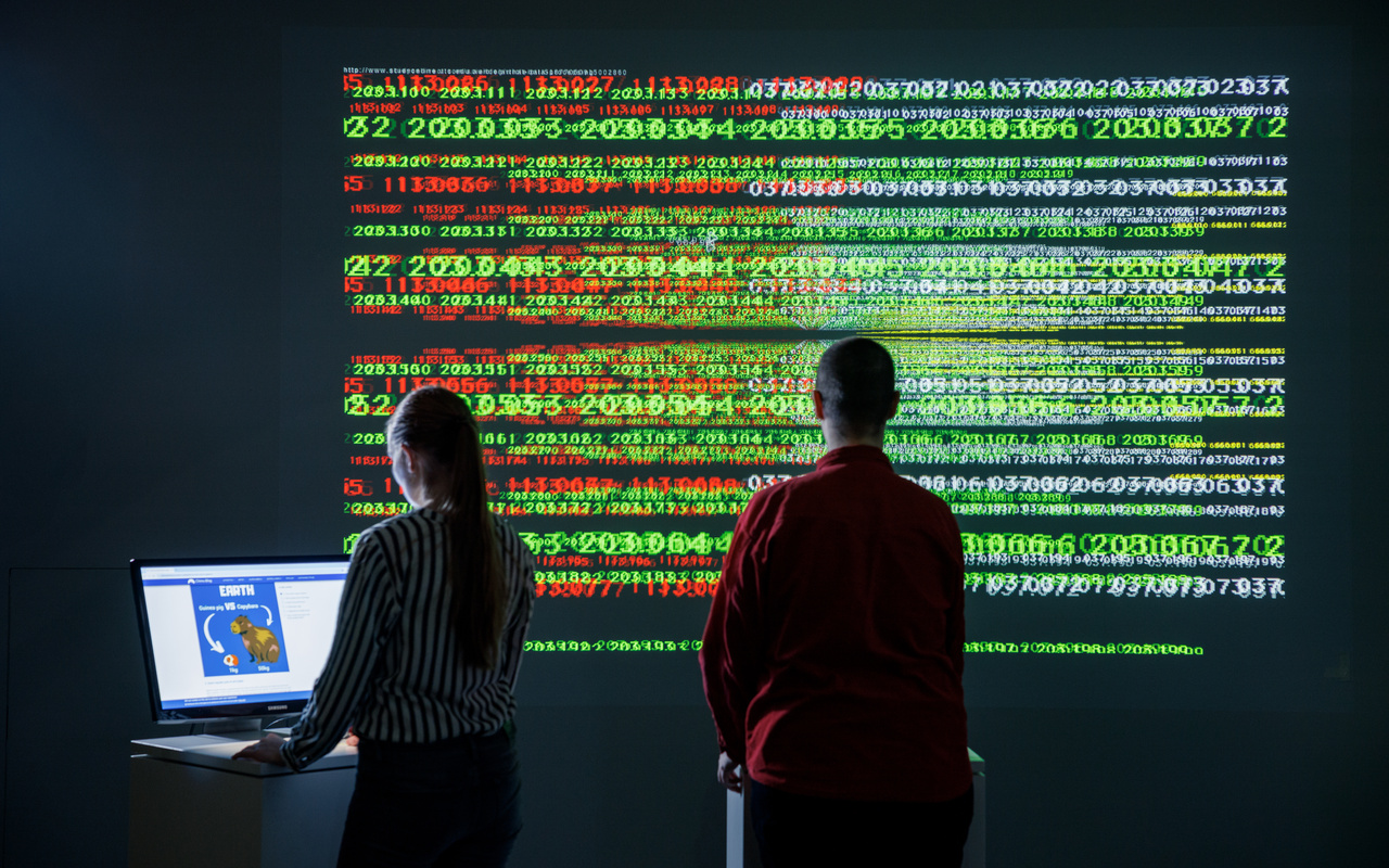 Eine Person steht in einem dunklen Raum vor einer Wand voller grüner und roter Ziffern. Links davon steht eine andere Person an einem Computerbildschirm.