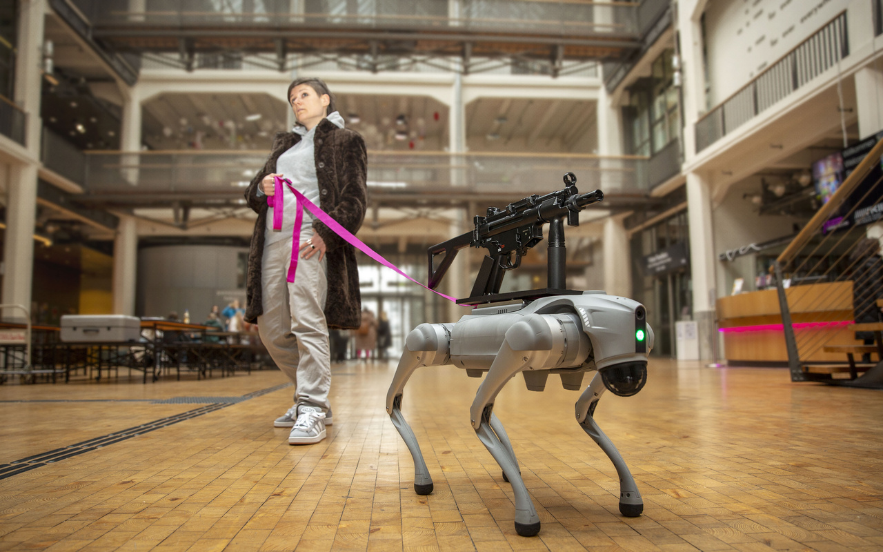 Ein Roboter Hund mit einem Sturmgewähr auf dem Rücken, der von einer pinken Leine von einer Person mit braunen Haaren und Pelzmantel gehalten wird.
