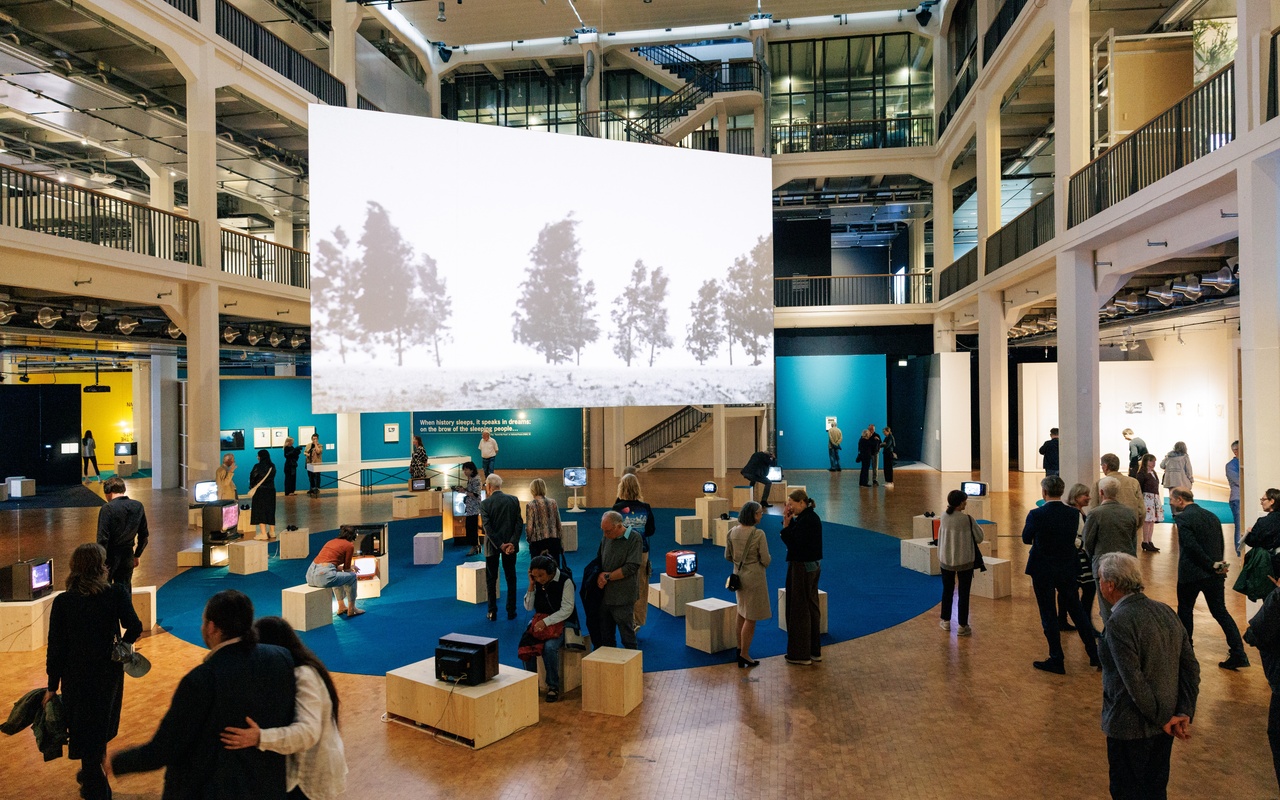 Besuchende betrachten eine multimediale Ausstellung mit Vintage-Fernsehern und großformatiger Videoprojektion in einem offenen Museumsraum.