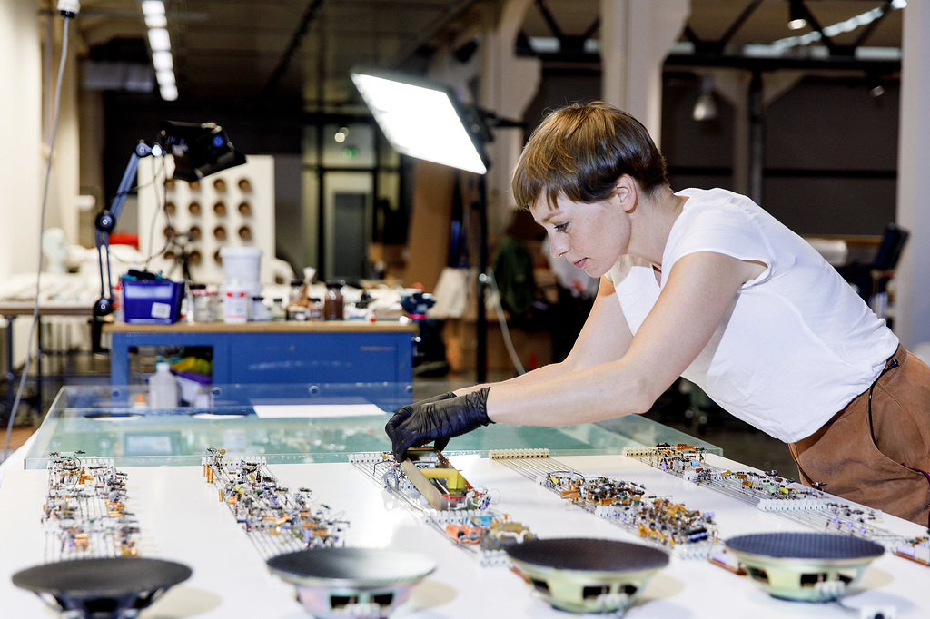 A large room can be seen, in the middle of which is a desk with many electronic objects. A woman with short hair and gloves bends over the table.