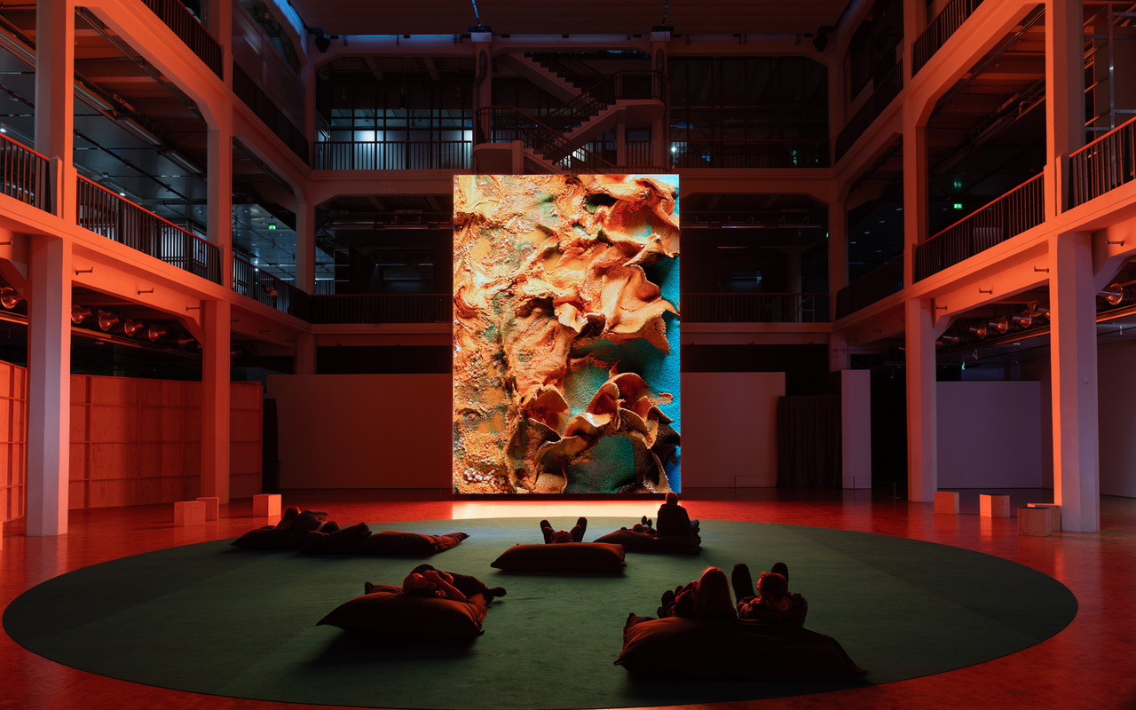 Personen sitzen vor einem riesigen Bildschirm, auf dem ein abstraktes Kunstwerk zu sehen ist.