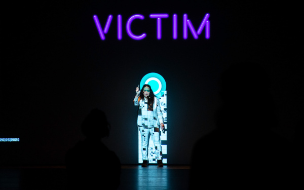Noa Frenkel stands in front of a background. On this background you can read »Victim« in large letters. Projected behind and on her is a rectangle with a circle on its top.