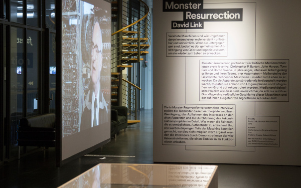 Blick in die Ausstellung »Monster Resurrection«: Eine Projektion, eine Wand mit Beschriftung, eine Vitrine