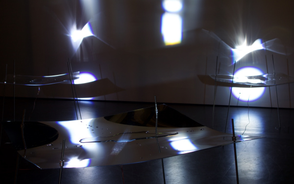 Ein dunkler Raum mit zarten Strukturen aus Plexiglas und Flüssigkeit, auf die Licht fällt.