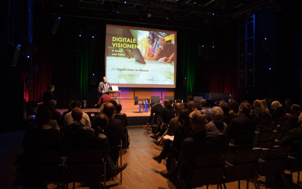 A room filled with people and a man on stage, giving a lecture.