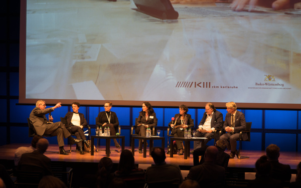 Seven people sit in a semicircle on a stage and discuss with eachother.