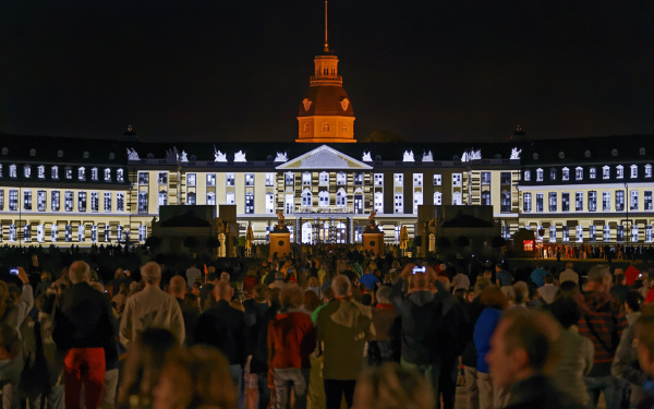 Menschen stehen vor dem Karlsruher Schloss, auf das Figuren projiziert wird