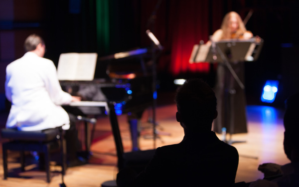 A man in a white suite playing piano. A woman in a black dress playing violin.