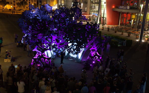 A black construction dipped in purple light. In the midst of the construction people are standing.
