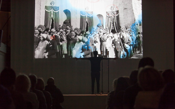 A man stands with spread arms in front of a screen on which people can be seen