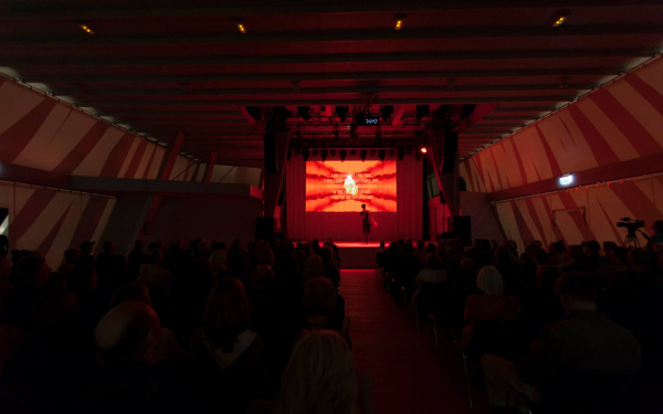 A man in front of a red screen. In front of it there are sitting many people.