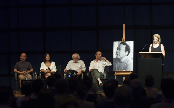 Vier Personen sitzen auf der Bühne. Am Rednerpult spricht eine Frau und ein großes schwarz-weiß Foto von einem jungen Mann ist aufgestellt.
