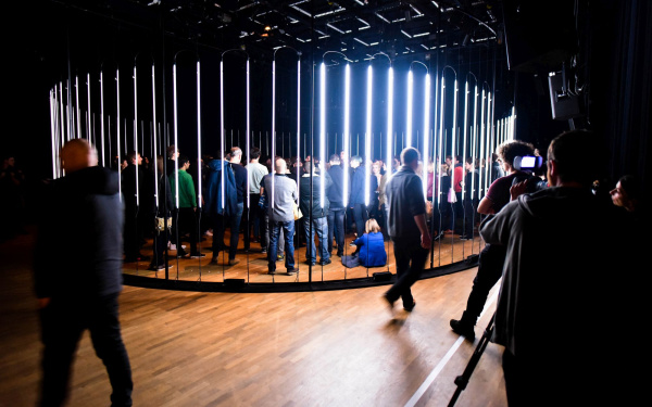 A circle of glowing neon tubes, many people standing in it