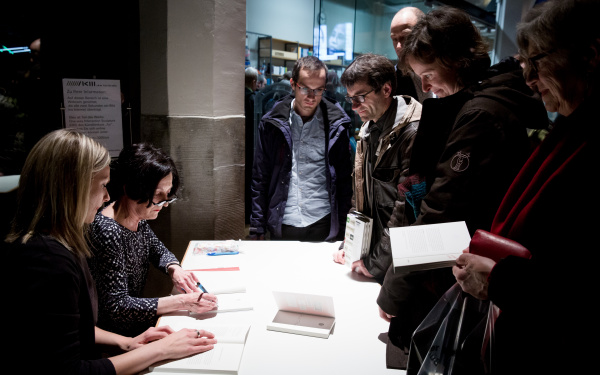 People standing in front of a desk. A woman sitting at the desk signing books