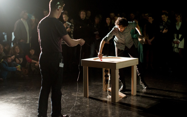 Two men. One holds a string, which is attached to a table. The other circles with a stiletto in a table