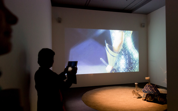 A woman taking a photo of a canvas on which the hand of a woman is be seen. On the floor of a small car with high heels made of cardboard off