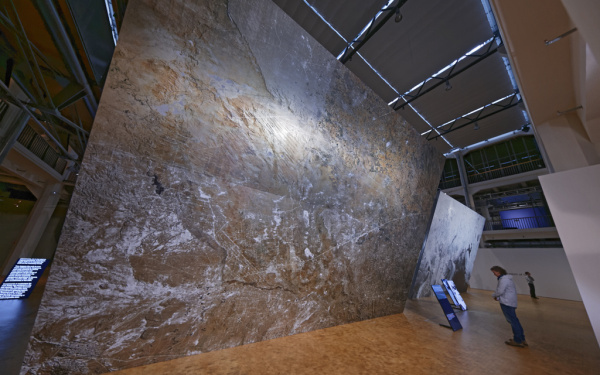 A person standing in front of a huge canvas on which a total of a cracked ground can be seen