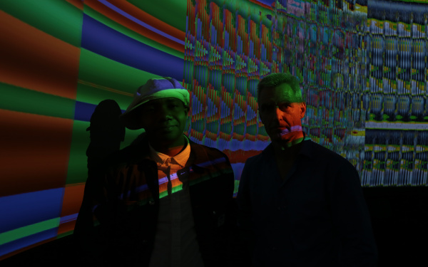 Two men standing in a dark room in front of a screen with colorful colors.