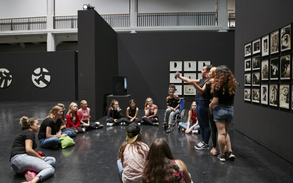 A group of girls sitting in an exhibition space, while three of them are taking a selfie in front of an artwork.