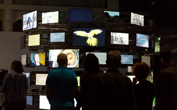 Das Bild zeigt BesucherInnen vor einer kreisförmig angeordneten Vielzahl von unterschiedlich bespielten Bildschirmen. 