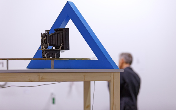Das Bild zeigt eine Kamera, die durch ein hohles, blaues Dreieck gerichtet ist. 