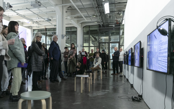 The photo shows the museum balcony of the ZKM with the exhibition of the conference. On the right there are three screens with headphones and opposite are many people.