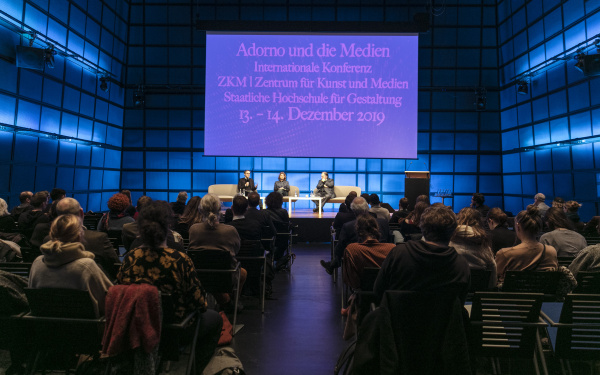 Im abgedunkelten Medientheater erleuchtet das Plakat der Adorno und die Medien Konferenz. Auf dem Podium sitzen von links nach rechts Johan Hartel, Loudmila Voropai und Peter Weibel