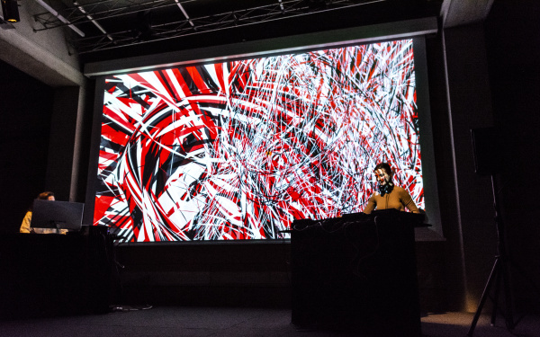 Zu sehen ist eine Visual-Sound-Performance. Zwei Frauen stehen an den unteren Ecken des großen Bildschirmes an ihren Soundcontrollern während im Hintergrund eine Farbstreifen aus rot schwarz und weiß eineinander verschmelzen.
