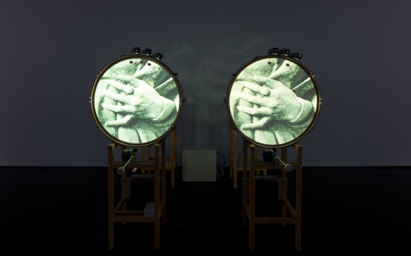 Zu sehen sind zwei stehende Trommeln, welche jeweils ein Bild von zwei ineinander gefalteten Händen auf der runden Fläche zeigen. 